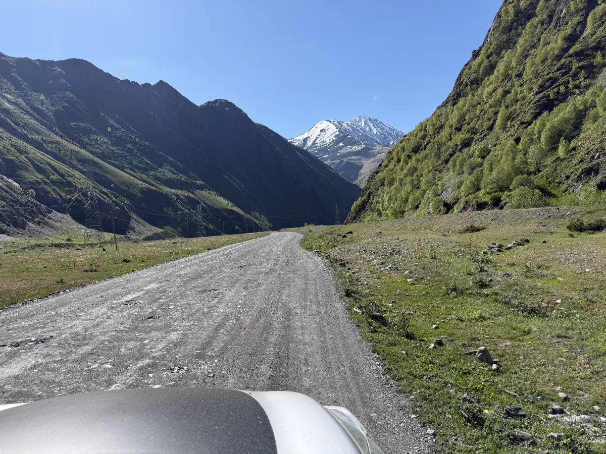 Wide compacted gravel road heading into a narrowing valley with a snow-capped peak ahead