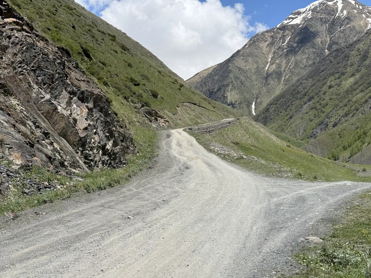 Gravel road bending beneath dark rock crags with a stone retaining wall ahead