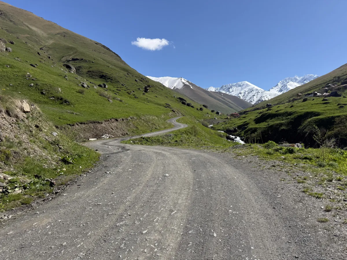 Gravel road descending in S-curves towards a mountain stream with snowy peaks beyond