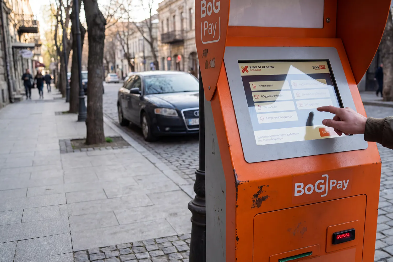 A bright orange Bank of Georgia 'Bog Pay' payment terminal on a Tbilisi sidewalk, with a person's hand using the touchscreen to pay for street parking.